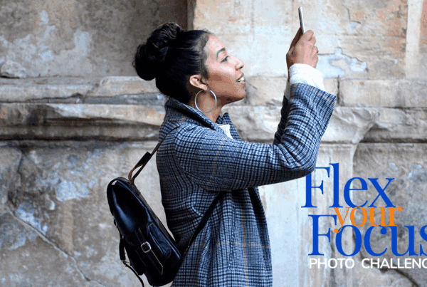 woman with dark hair wears a black backpack and a blue blazer jacket while she holds up her phone to take a photo