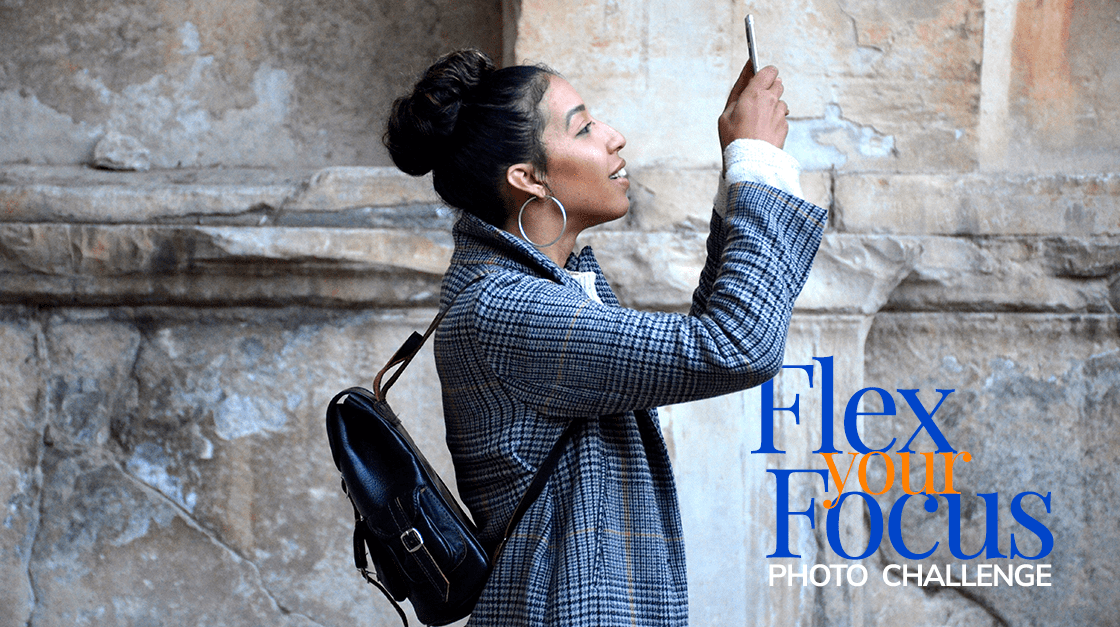 woman with dark hair wears a black backpack and a blue blazer jacket while she holds up her phone to take a photo